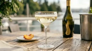 A photorealistic 4K close-up of a classic, wide-mouthed coupe glass filled with Prosecco, featuring a delicate gold rim. The glass is set on the weathered wooden table of a sunlit Vancouver waterfront patio, with the Bottega bottle subtly blurred in the background.